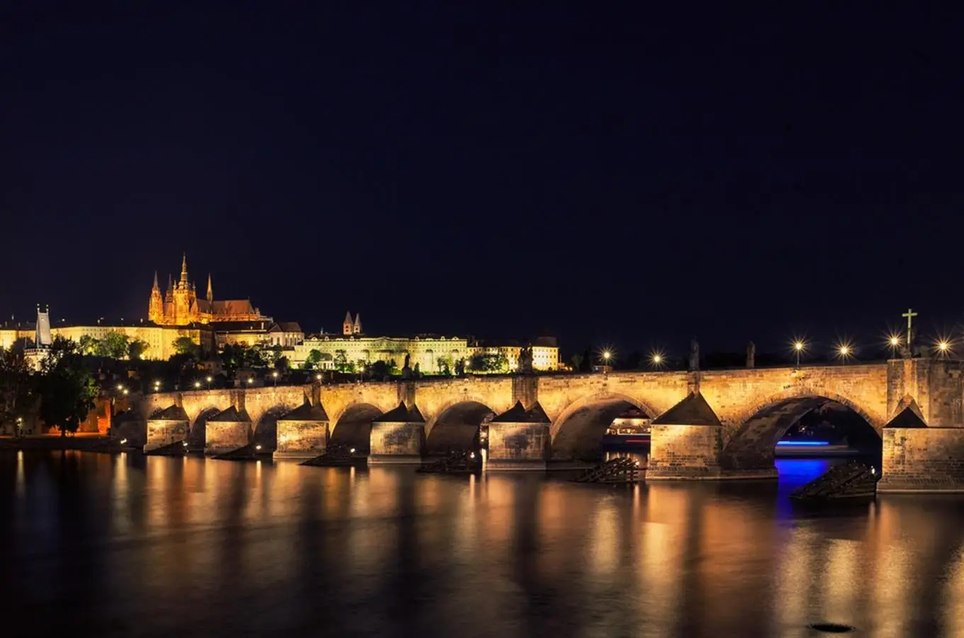 Noční Karlův most s lucernami, odrazy světel na tmavé hladině Vltavy